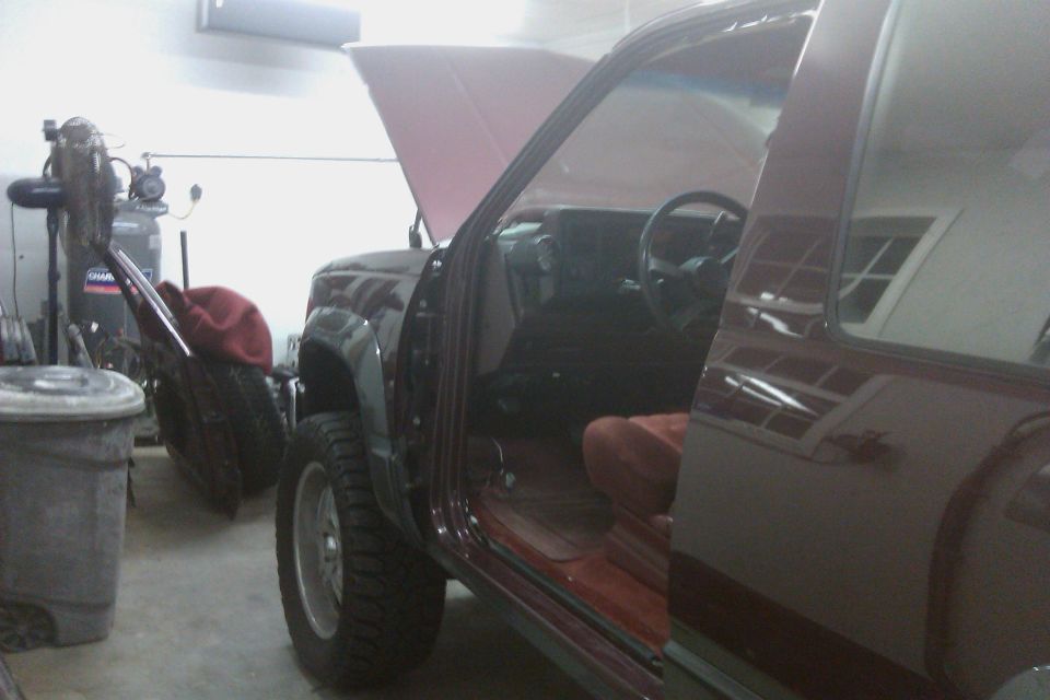 Maroon SUV interior with open door, showing dashboard and seats. Hood is up. Tires and trash can visible