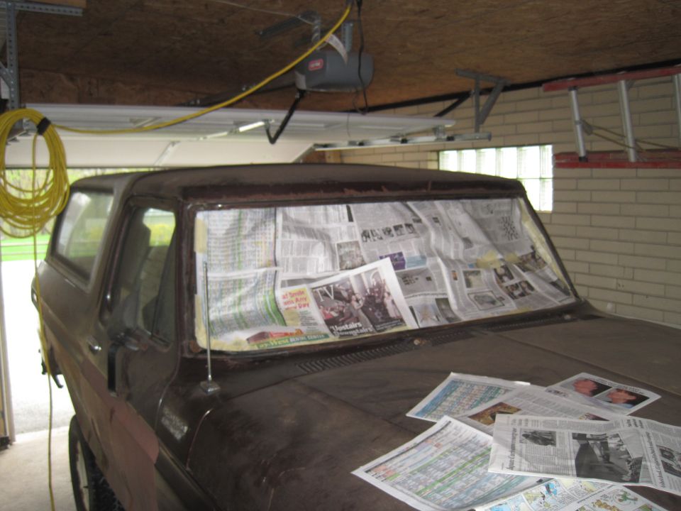 Old SUV in garage, windshield covered in newspapers. Some newspapers are on the hood too
