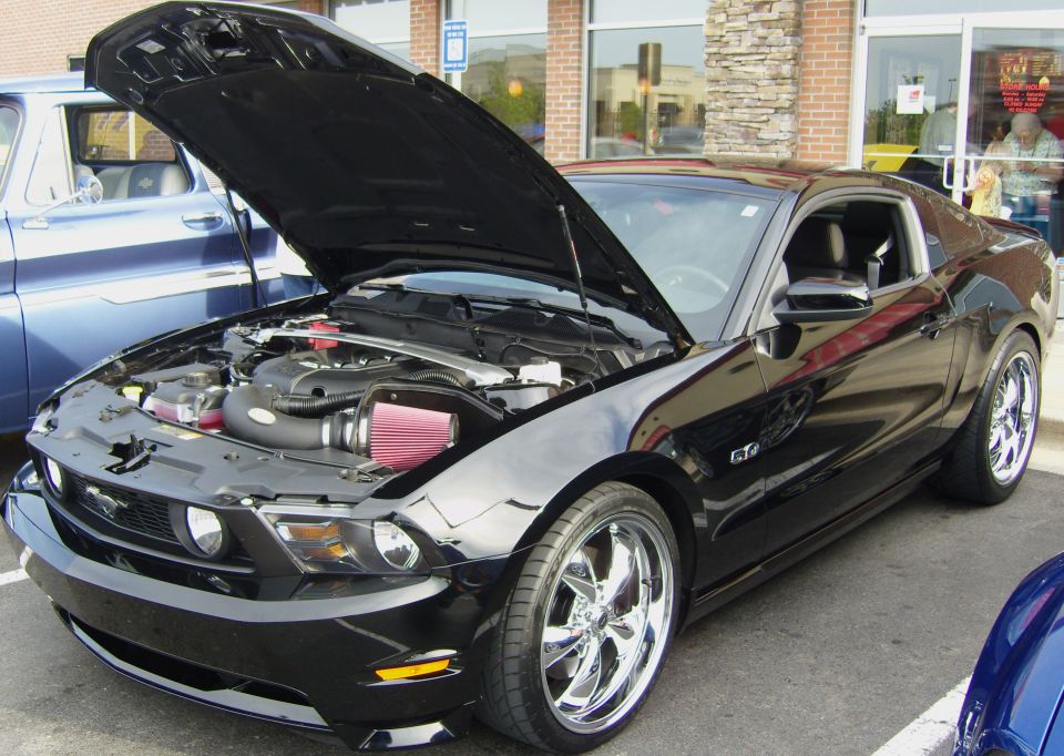 Black Ford Mustang GT with hood open, showing engine and aftermarket air intake. Chrome wheels and GT badge visible