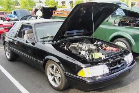 2007 Ford Mustang - Black Fox-body Ford Mustang LX notchback at an outdoor car show with the hood propped open, showing a 5.0 V8 with aftermarket intake and re…