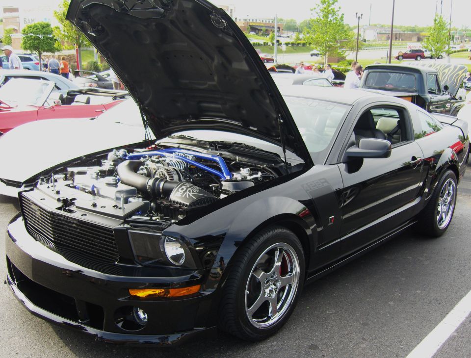 Black Roush Mustang at a car show, hood up showing supercharger with blue intake piping