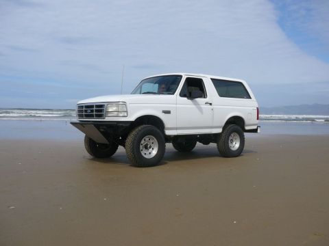 1988 Ford Bronco - White 1995 Ford Bronco with a custom front bumper parked on a sandy beach with ocean waves in the background.