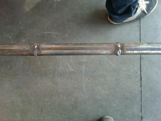 Close-up of a welded exhaust pipe section on a concrete floor. A person's legs and shoes are visible at the top