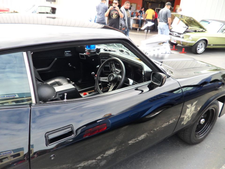 Black coupe interior visible, side view of car with aftermarket wheels. Another car with hood open in background