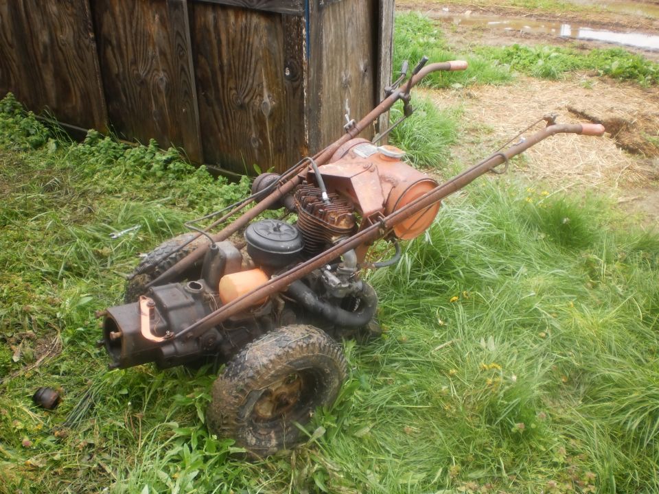 Rusty walk-behind tiller with orange gas tank and engine, sitting in tall grass next to a wooden structure