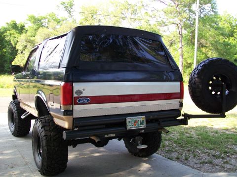 1990 Ford Bronco - This lifted Ford Bronco features a soft top and a rear-mounted spare tire, sitting on a paved surface.
