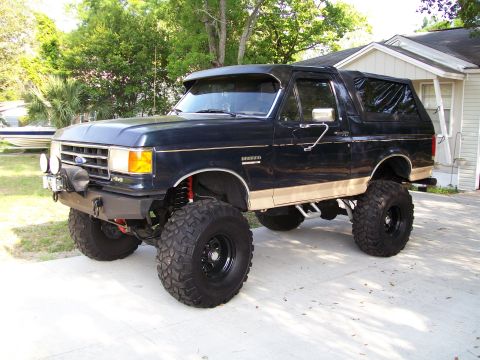 1990 Ford Bronco - This lifted 1990 Ford Bronco features a dark blue exterior with tan lower body trim and prominent off-road tires.