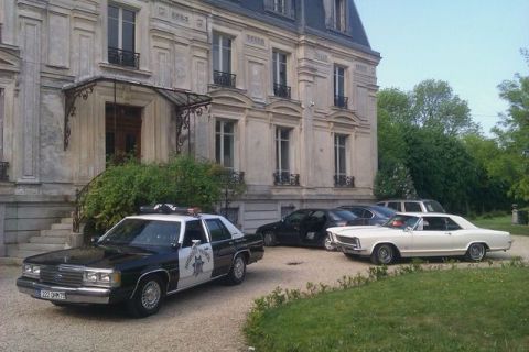 1991 Ford Crown Victoria - A police car and a white coupe are parked in front of a large building with other vehicles in the background.