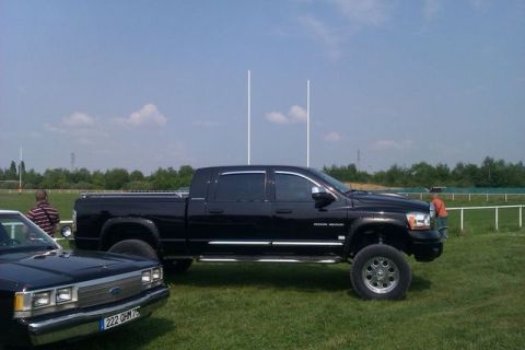 1991 Ford Crown Victoria - A black Dodge Ram pickup truck is parked on a grassy field next to a dark sedan under a blue sky.