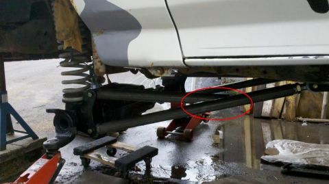 1996 Ford Bronco - Low ground-level view under the passenger side of a white lifted truck on jack stands and a red floor jack, showing the front suspension wi…
