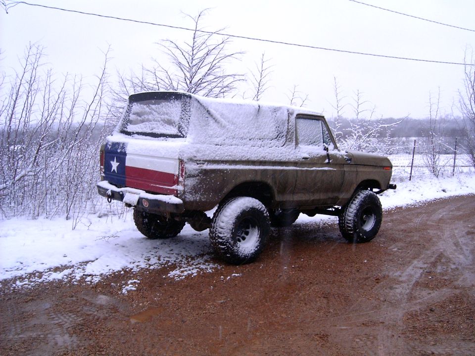 Snow-covered olive Ford Bronco with Texas flag tailgate on a red dirt road