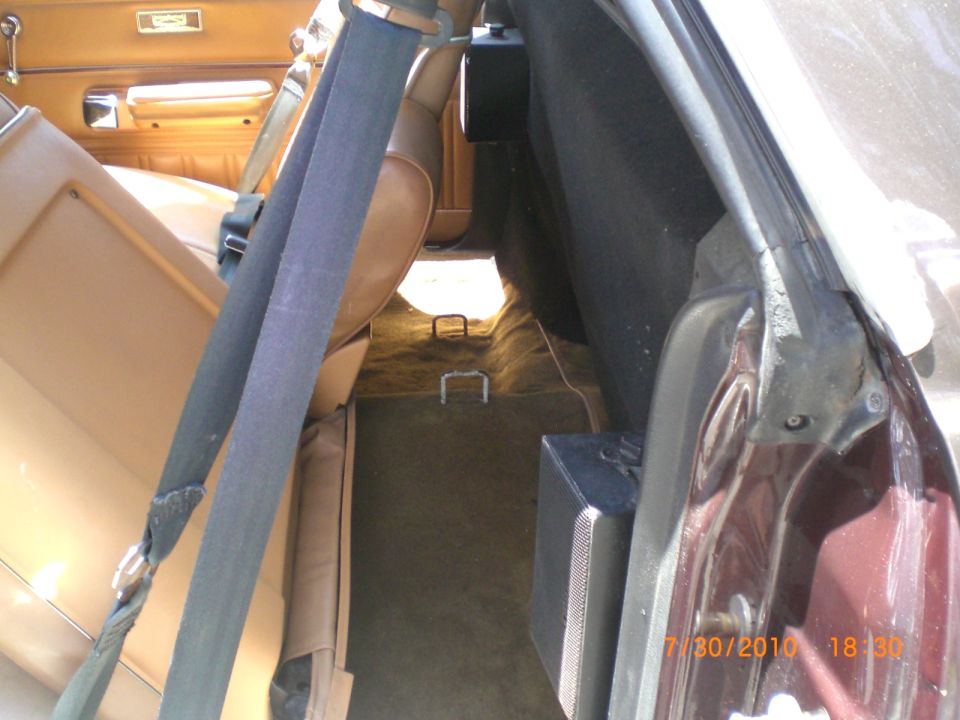 View of the rear interior of a vehicle, showing tan seats, seatbelts, and a carpeted cargo area with speakers