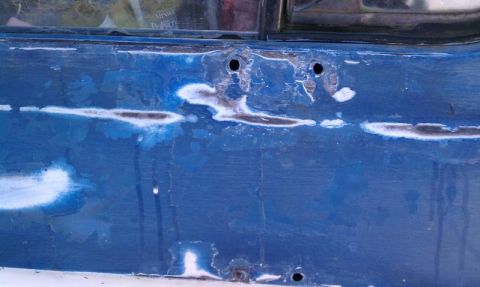 1979 Ford Bronco - Detail of a blue vehicle's bodywork showing areas of rust and primer beneath the paint.