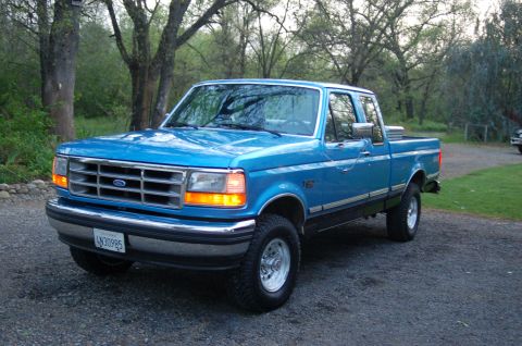 1992 Ford F-150 - A blue 1992 Ford F-150 pickup truck is parked outside on a gravel driveway with trees in the background.