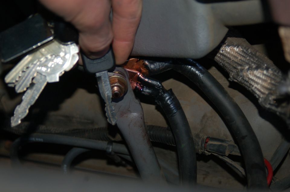 Hand holding keys near a corroded electrical battery cable connection under a vehicle