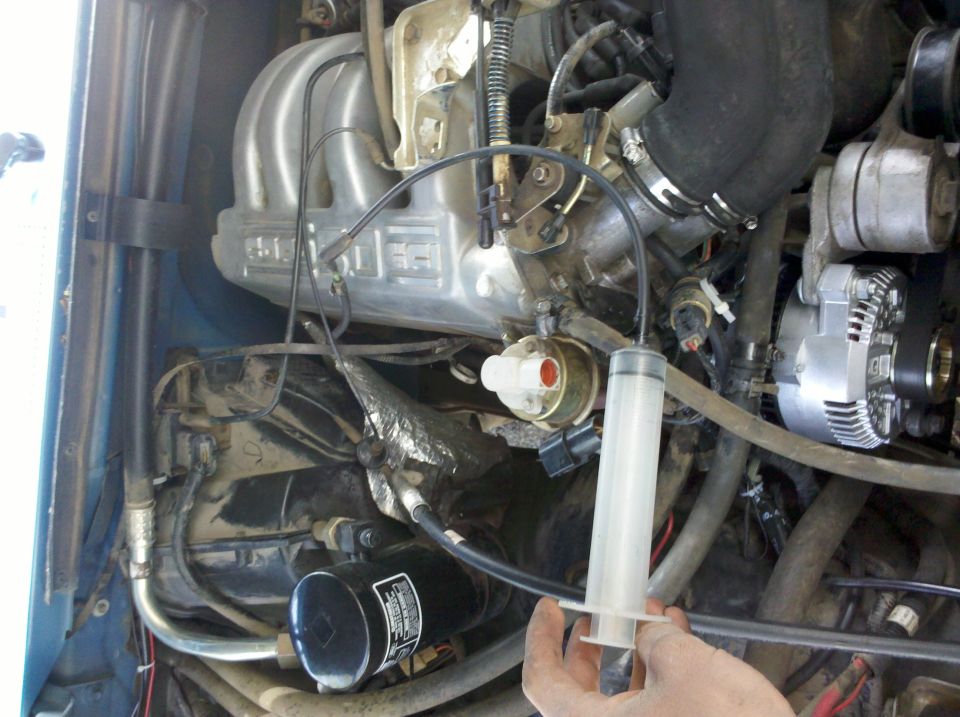 1992 Ford F-150 engine bay, close-up on the intake manifold and alternator, with a syringe in hand