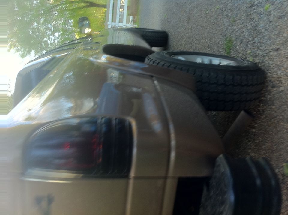 Rotated blurry photo of a tan pickup rear corner with tinted taillight and a loose wheel on gravel