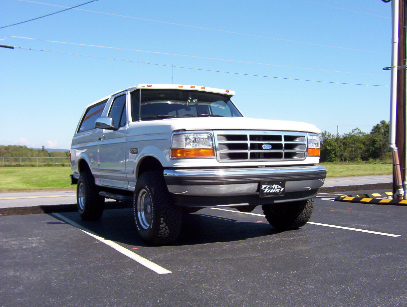 Lifted white Ford Bronco SUV with 'FEAR THIS!' front plate parked in a lot under blue sky