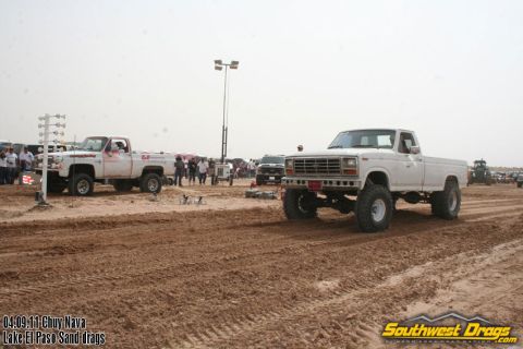 1995 Ford Bronco - A lifted white pickup truck competes in a sand drag race, with another truck and spectators in the background.