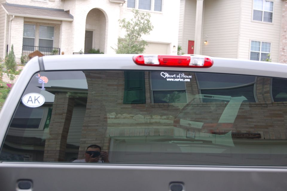 Gray truck rear window with AK and Heart of Praise stickers. Reflection shows a house and a person with a camera