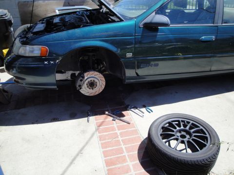 1995 Ford Taurus - A green 1995 Ford Taurus is undergoing maintenance, with its hood up and front wheel removed to access the braking system.