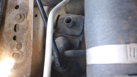 1994 Ford Bronco - Tight underhood close-up of a small dark reservoir or valve with an open filler neck, tucked between a rusty stamped-steel bracket on the l…