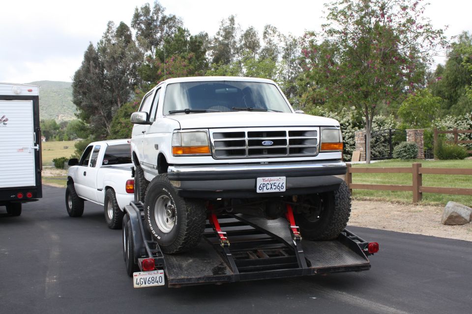 Lifted white Ford Bronco on a black car hauler trailer, secured with red straps, California plate 6PCX566