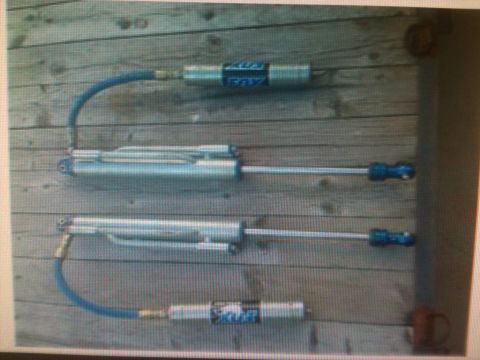 1992 Ford Bronco - Three Fox shock absorbers are laid out on a wooden surface, showing their silver bodies and blue accents.