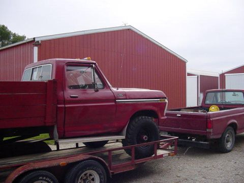 1992 Ford Bronco - A red 1977 Ford F-250 flatbed truck is loaded onto a trailer, with another red pickup truck visible behind it.