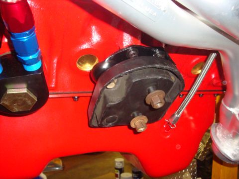 1989 Ford Bronco - A close-up view of a red engine block shows a black motor mount secured with bolts, alongside part of a metal exhaust pipe.