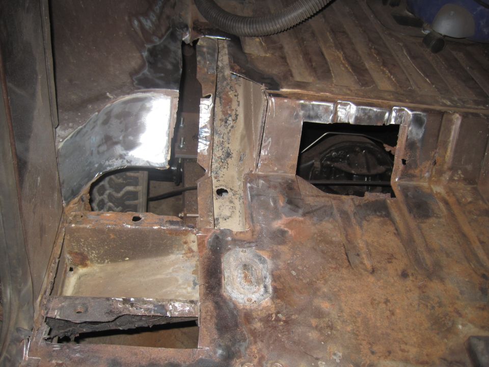 Underside of vehicle showing rusted floor pan and metal repairs. Tire visible in wheel well