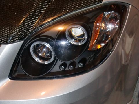 2009 Chevrolet Cobalt - Detail of a vehicle's headlight assembly, projector-style low beams and an integrated turn signal. The carbon fiber hood is partially visib…