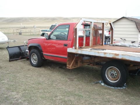 1994 GMC Sierra 3500 - A red flatbed truck equipped with a snow plow is parked on a grassy area next to a building.