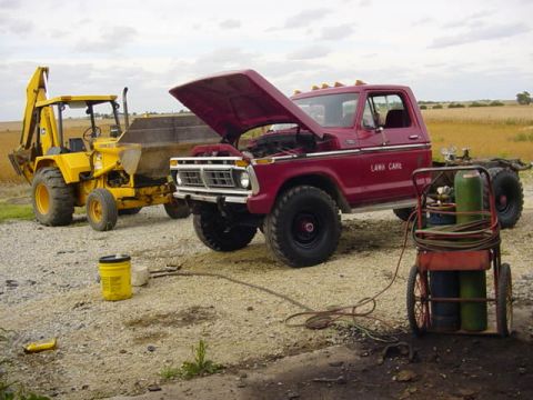 1992 Ford Bronco - A red pickup truck, possibly from the 1970s, is parked next to a yellow backhoe with welding equipment nearby.