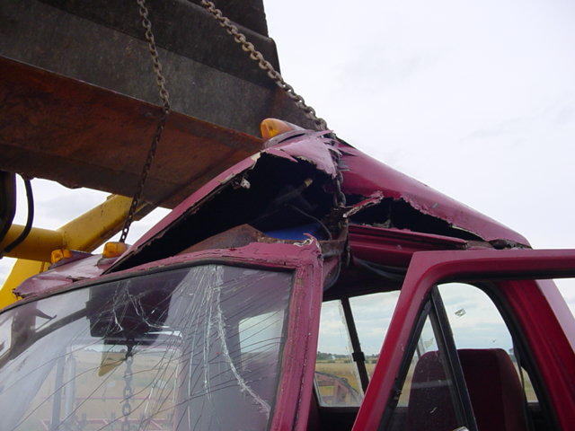 Red truck cab with crushed roof and shattered windshield being lifted by a chained loader