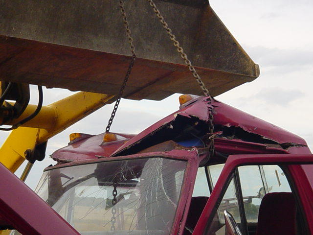 Damaged red pickup truck cab being lifted by a yellow excavator bucket and chains