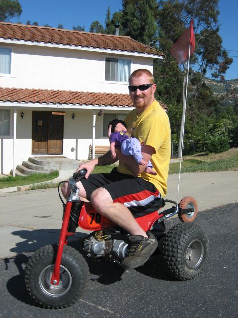 1900 Honda Big Red - Man with baby on a red three-wheeled ATV, with a house and trees in the background.