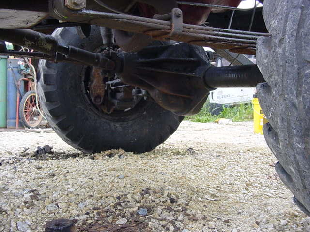 Rear axle and leaf springs of a large vehicle, with a partial view of a tire and gravel ground