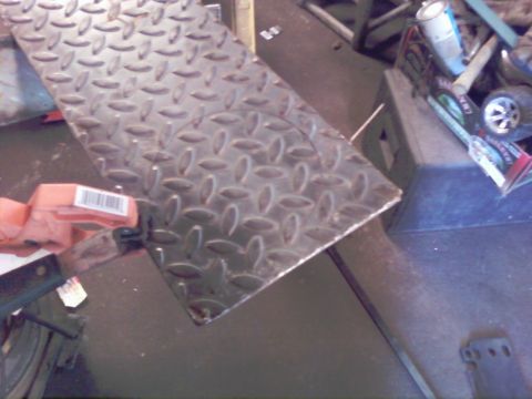 1989 Ford Bronco - A close-up of diamond plate metal with a circular cut, held by an orange clamp, in a workshop setting.
