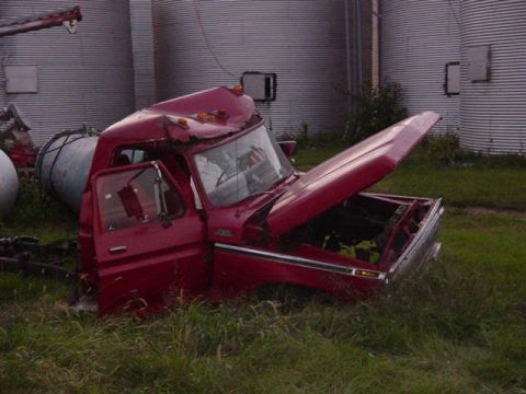 1992 Ford Bronco - A damaged red truck cab sits in a field, its hood and door open, with large silos in the background.