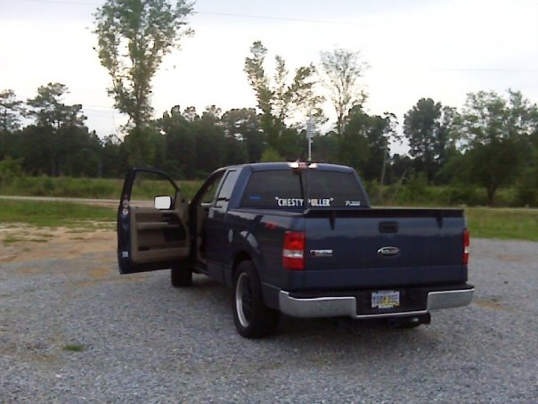Dark blue Ford F-150 pickup from rear three-quarter view with driver doors open, parked on gravel