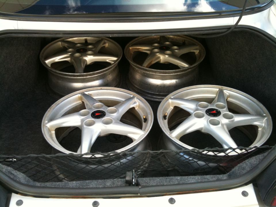 Four Pontiac 5-spoke alloy wheels without tires stored in the trunk of a white sedan