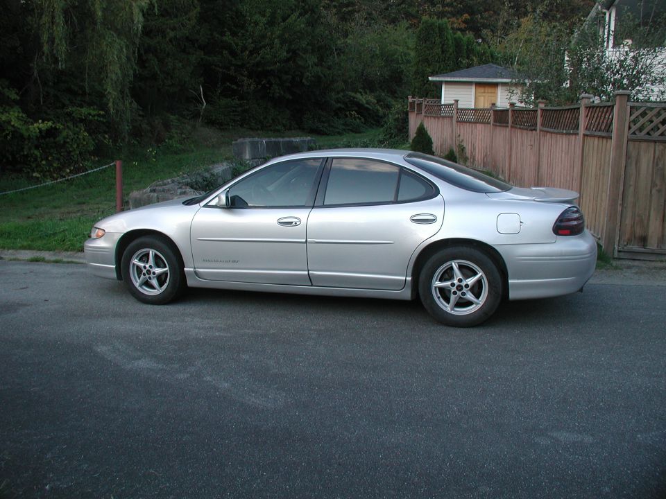 Silver Pontiac Grand Prix sedan, driver side view, parked by a wooden fence and trees