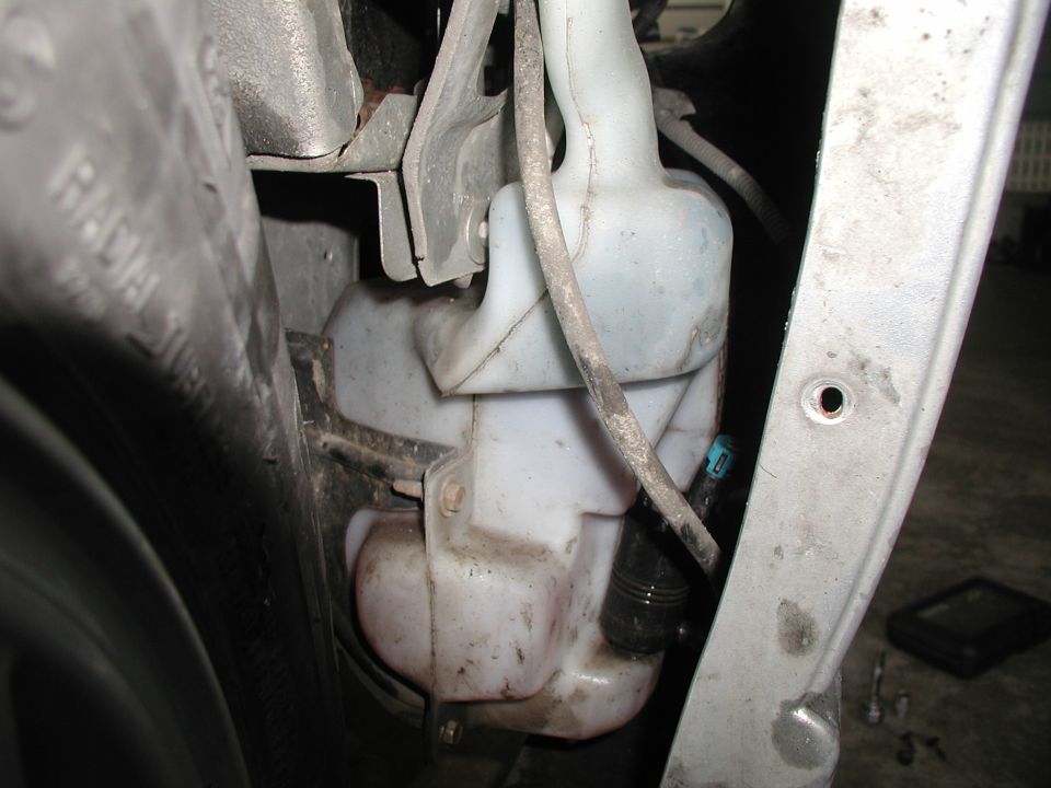 White washer fluid reservoir tank in engine bay near wheel well with hoses and bracket