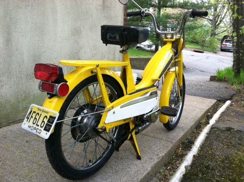 1981 Garelli Rally Sport - A yellow moped is parked on a sidewalk next to a concrete wall, with a street in the background.