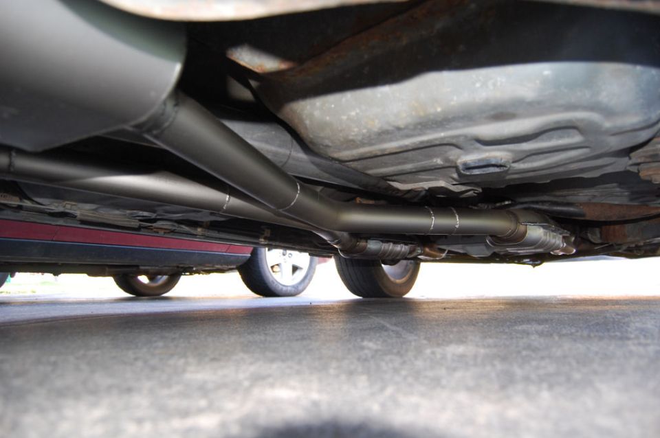 Underside of a car showing a custom exhaust system with two pipes and a catalytic converter, with another car visible underneath