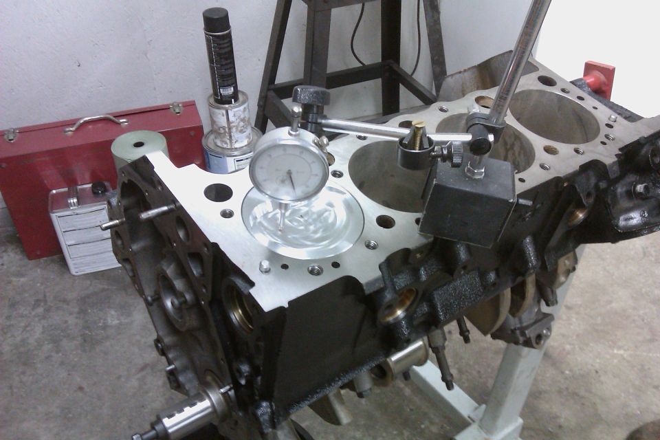 Engine block on stand with dial indicator measuring cylinder bore. Red toolbox and spray can in background