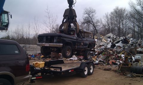 1978 Ford Bronco - Junkyard crane lifts a 1978 Ford Bronco, with a trailer and another SUV present.