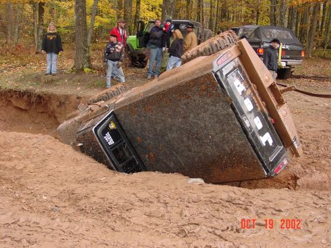 1986 Jeep J-10 - A truck is stuck and overturned in a muddy pit, with onlookers observing the scene.