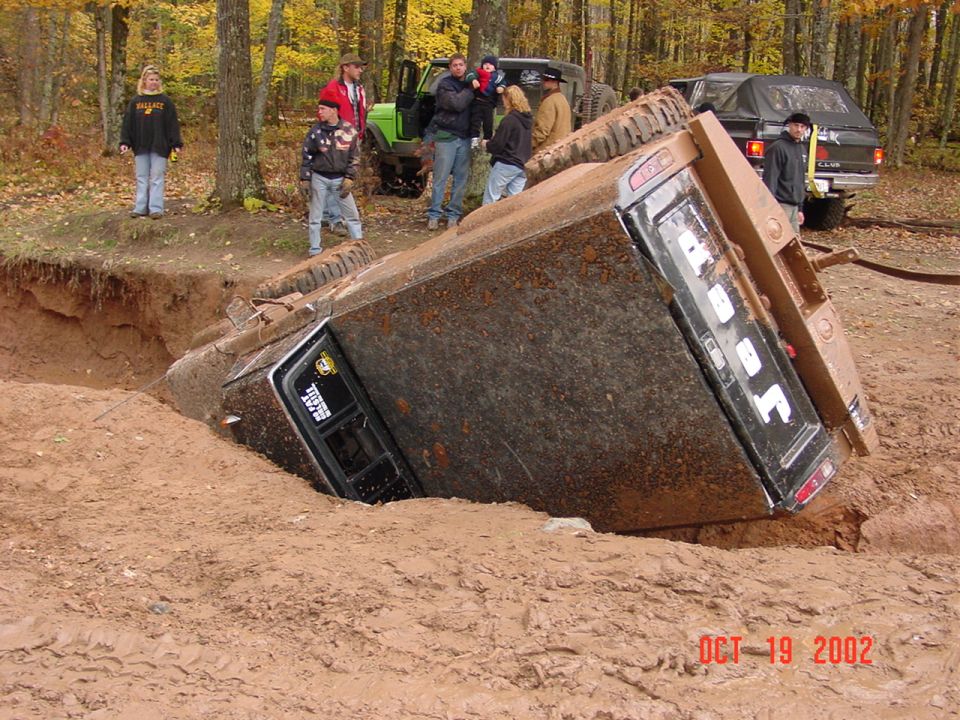 Mud-covered truck overturned in a dirt pit, with people watching